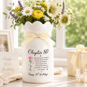 May include: A white ceramic vase with a scalloped rim and a cream-colored ribbon tied around the neck. The vase features black text reading Chapter 80, a poem, and Happy 80th Birthday, alongside a pink flower illustration and small butterflies.