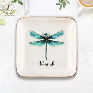 May include: A white ceramic trinket tray with a gold rim features a blue dragonfly with a black outline and the name "Hannah" written in black cursive.