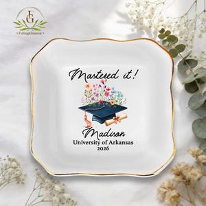 May include: White ceramic trinket dish with a gold rim. The dish features a graduation cap design with the text "Mastered it!" and the name "Madison" with "University of Arkansas 2026".