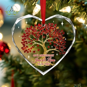 May include: A heart-shaped, clear glass ornament with a red ribbon. Inside, a golden tree with red leaves is depicted, along with the text "40th Anniversary" and names "Jane & Liam" with the date "10-18-1985".