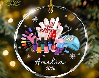 May include: A round, clear glass ornament featuring a graphic of a hand applying red nail polish, surrounded by various colorful nail polish bottles, a nail color display wheel, a blue UV lamp, and white snowflakes, with the text Amelia 2026.