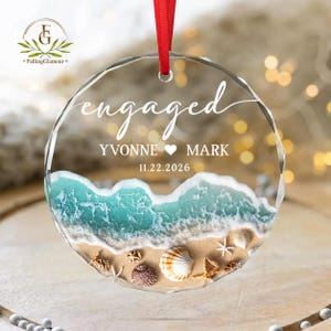 May include: A clear, round ornament with the word "engaged" in elegant script. The ornament features a beach scene with turquoise waves, sand, and seashells. The names "YVONNE ♥ MARK" and the date "11.22.2026" are also displayed. A red ribbon is attached for hanging.