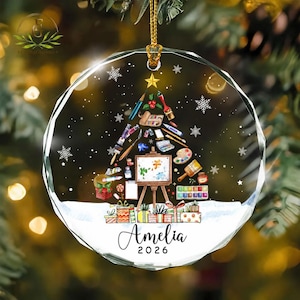 May include: A circular clear glass Christmas ornament featuring a printed design of an artist-themed Christmas tree made of paintbrushes, palettes, and tubes, topped with a gold star. The base shows wrapped gifts and the name Amelia 2026.