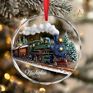 May include: A clear, round Christmas ornament with a detailed illustration of a vintage train. The train is blue, green, and red, with the name "Nicholas" and the year "2025" printed on the front. A red ribbon is attached for hanging.