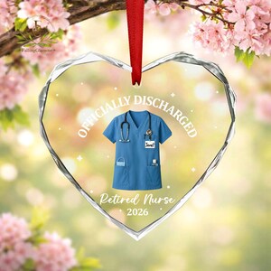K&ouml;nnte beinhalten: Herzf&ouml;rmiger Glasornament mit rotem Band. Der Ornament zeigt ein blaues medizinisches Oberteil mit einem Stethoskop, den Text "OFFICIALLY DISCHARGED", "Retired Nurse" und das Jahr "2026". Ein Namensschild lautet "Janet".