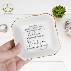 May include: White ceramic trinket dish with a gold rim, featuring the text "There are people in this world who make things better wherever they go. Thank you for being one of them, Alice." A ring and a small clock are visible.