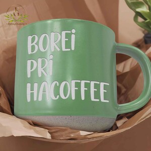 May include: A green ceramic coffee mug with the words "BOREI PRI HACOFFEE" in white lettering. The mug has a grey base and a curved handle. The mug is sitting in a brown paper bag.