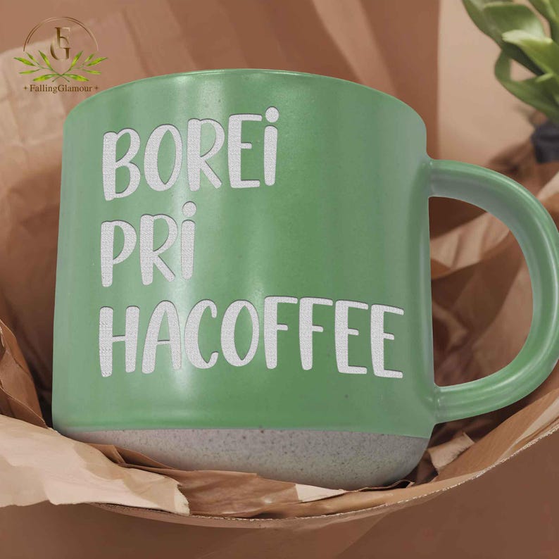 May include: A green ceramic coffee mug with the words "BOREI PRI HACOFFEE" in white lettering. The mug has a grey base and a curved handle. The mug is sitting in a brown paper bag.
