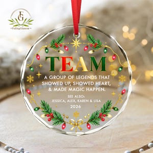 May include: A circular clear glass Christmas ornament with a red ribbon hanger. It features a green and red wreath design with Christmas lights, snowflakes, and the text: TEAM A GROUP OF LEGENDS THAT SHOWED UP, SHOWED HEART, & MADE MAGIC HAPPEN. SEE ALSO: JESSICA, ALEX, KAREN & LISA 2026.
