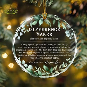 May include: A clear, round ornament with the text "DIFFERENCE MAKER" and a definition. The ornament is decorated with green leaves and has a gold string for hanging. The name "Amanda" is also included.
