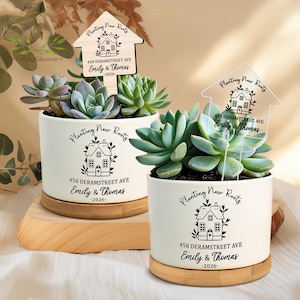 May include: Two white cylindrical ceramic planters with bamboo bases, each containing a green succulent. The planters feature black text reading Planting New Roots, 458 Deramstreet Ave, Emily & Thomas, 2026, and a house illustration.