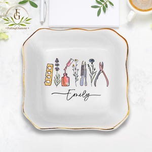 May include: White ceramic trinket dish with a gold rim, featuring a watercolour design of nail care tools and a nail polish bottle. The word "Emily" is written in cursive script below the design.