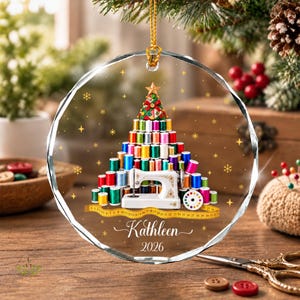 May include: A circular glass ornament featuring a Christmas tree constructed from colourful sewing thread spools, topped with a star. A white sewing machine sits at the base with a yellow measuring tape, the name Kathleen, and the year 2026 printed below.