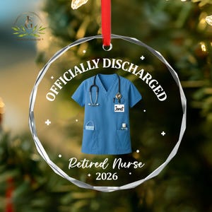 K&ouml;nnte beinhalten: Ein klarer, runder Weihnachtsschmuck mit den Worten "OFFICIALLY DISCHARGED" und "Retired Nurse 2026" in Wei&szlig;. Der Schmuck zeigt einen blauen medizinischen Kittel mit Stethoskop, Gesichtsmaske und H&auml;ndedesinfektionsmittel. Ein rotes Band ist zum Aufh&auml;ngen befestigt.