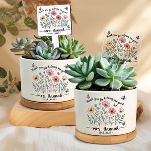 May include: Two white ceramic succulent planters with wooden bases. Each planter has a floral design and the text "Thank you for helping me grow" and "Mrs. Hannah Love Julia". The planters contain succulents and a sign.
