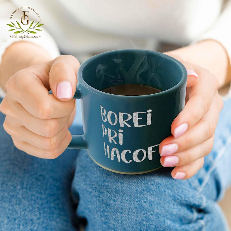 May include: A teal ceramic mug with the words "BOREI PRI HACOF" in white lettering. The mug contains a dark liquid, possibly coffee or tea, and steam is rising from it. The mug is held by a person.