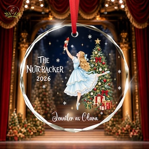 Peut inclure: Une décoration en verre facetté ronde avec un ruban rouge, illustrant une ballerine en robe bleu clair tenant un casse-noisette. Le motif comprend un sapin de Noël décoré, le texte The Nutcracker 2026 et Jennifer as Clara.