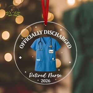 K&ouml;nnte beinhalten: Ein klarer, runder Weihnachtsschmuck mit rotem Band. Der Schmuck zeigt einen blauen medizinischen Kittel mit Stethoskop, Gesichtsmaske und kleiner Flasche. Der Text auf dem Schmuck lautet "OFFICIALLY DISCHARGED", "Retired Nurse" und "2026".