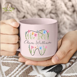 May include: A light pink ceramic mug with a floral tooth design and the name "Clara William". The design includes colorful flowers and green leaves. The mug has a light brown base and is held by a person.
