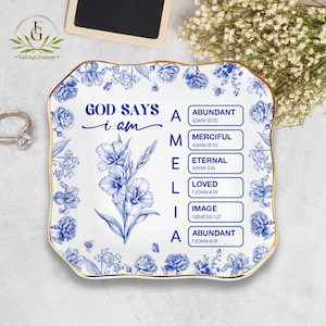 May include: A white ceramic decorative plate with a blue floral design and gold trim. The plate features the text "GOD SAYS i am" and the word "AMELIA" with associated biblical verses. The plate is square with a scalloped edge.