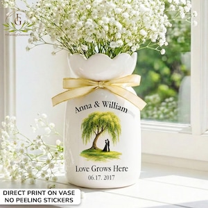 May include: A white ceramic vase with a scalloped top, tied with a gold ribbon, and filled with white flowers. The vase features a tree illustration and the text "Anna & William", "Love Grows Here", and the date 06.17.2017.