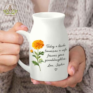 May include: White ceramic mug with a handle, featuring an orange flower illustration and the text "Today a bride tomorrow a wife forever your granddaughter. Love, Sophia" with a heart symbol. The mug is held by a person.