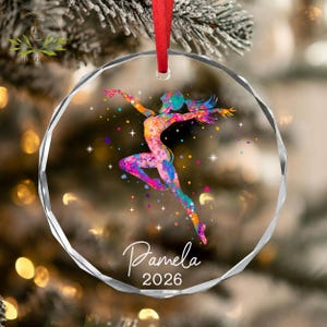 May include: A circular, faceted glass ornament featuring a colorful, watercolor-style silhouette of a gymnast in a leaping pose. The design includes small star accents and the printed text "Pamela" above the year "2026". A red ribbon is attached.