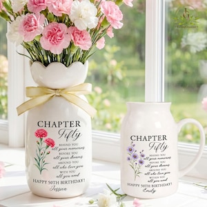 May include: A white ceramic vase with a scalloped rim and a cream ribbon, filled with pink carnations, alongside a white ceramic mug. Both items feature the text "CHAPTER Fifty" with floral illustrations and "HAPPY 50TH BIRTHDAY" printed on them.
