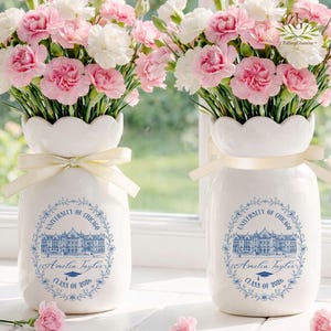 Pu&ograve; includere: Due vasi in ceramica bianca con bordi smerlati, ciascuno legato con un nastro color crema, contengono bouquet di garofani rosa e bianchi. I vasi presentano un disegno blu con il testo "UNIVERSITY OF CHICAGO", un nome e "CLASS OF 2026".