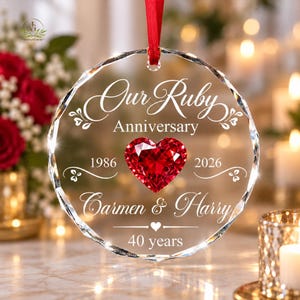 May include: A circular clear crystal ornament hanging from a red ribbon. The ornament features the engraved text Our Ruby Anniversary, 1986, 2026, Carmen & Harry, and 40 years, with a faceted red heart-shaped crystal centered in the middle.