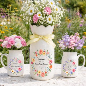 May include: A white ceramic vase and two matching mugs, each featuring a floral wreath design with the text Sister, EST. 1996, Maid of Honor, EST. 2026. The vase is tied with a yellow ribbon and holds a bouquet of white daisies and pink carnations.