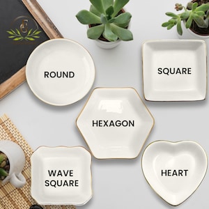 May include: A collection of white ceramic dishes in various shapes: round, square, hexagon, wave square, and heart. Each dish has a gold trim and a black label indicating its shape. The dishes are arranged on a white surface.
