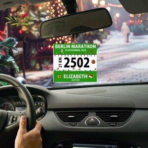 May include: A square hanging car ornament featuring a Berlin Marathon race bib design with the text BERLIN MARATHON, 10 NOVEMBER, 2025, 21 KM, 2502, ELIZABETH, and 2:27:23, decorated with Christmas lights, a snowman, a gift, and a Santa hat.