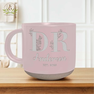 May include: A pink ceramic mug with a white floral design and the letters "DR" with the name "Anderson" and "EST. 2025" below.