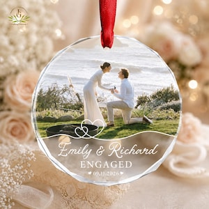 May include: A circular faceted glass ornament featuring a printed photograph of a marriage proposal on a beach. The ornament includes the white text "Emily & Richard", "ENGAGED", and the date "09.11.2026" with a red ribbon for hanging.