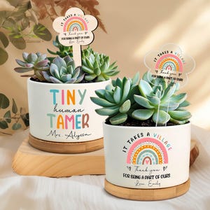 May include: Two white ceramic planters with succulents. One planter reads "TINY human TAMER Mrs. Alyssa" in colorful letters. The other planter has a sign that says "IT TAKES A VILLAGE Thank you FOR BEING A PART OF OURS Love Emily".