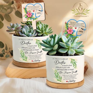 May include: Two white ceramic planters with succulents, each featuring a wooden sign with a stethoscope design and the name "Ashley." The planters have text that reads "A Truly Great Doctor" and a thank you message. The planters sit on wooden bases.