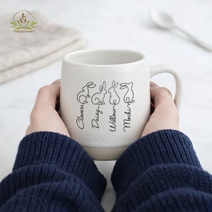 May include: A white ceramic mug with a speckled finish, featuring four line-drawn rabbits and names: Clover, Daisy, Willow, and Mochi. The mug has a curved handle and is held by a person wearing a navy blue sweater. The mug is a kitchen and dining product.