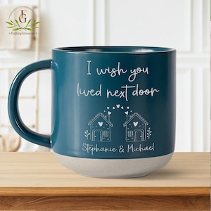 May include: Teal blue ceramic mug with a white handle. The mug has a white inscription that reads "I wish you lived next door" with two small houses and hearts above the text. The bottom of the mug is a light gray color and has the names "Stephanie & Michael" written in white.