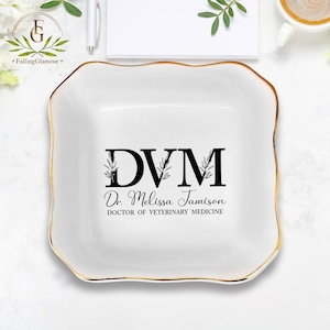 May include: White ceramic trinket tray with a gold rim. The tray features a black and white design with the text "DVM Dr. Melissa Jamison DOCTOR OF VETERINARY MEDICINE".