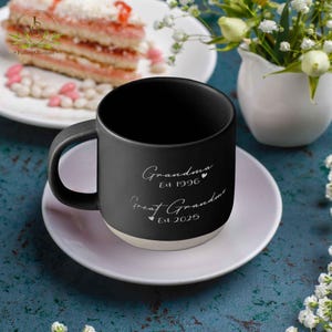 May include: Black ceramic coffee mug with a white inscription that reads "Grandma Est 1996" and "Great Grandma Est 2025" with a heart symbol between each line of text. The mug is sitting on a white saucer.