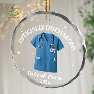K&ouml;nnte beinhalten: Ein klarer, facettierter Glasornament mit einem goldenen Faden zum Aufh&auml;ngen. Der Ornament zeigt ein blaues medizinisches Scrub-Top mit Stethoskop, Gesichtsmaske und H&auml;ndedesinfektionsmittel. Der Text lautet "OFFICIALLY DISCHARGED" und "Retired Nurse 2026".