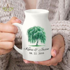 May include: White ceramic mug with a handle, featuring a green weeping willow tree design. The mug has the names "Katie & Jason" and the date "08. 22. 2016" printed on it. The mug is held by a person.