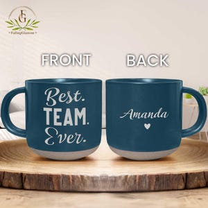 May include: A navy blue ceramic mug with a grey base. The front of the mug reads "Best. TEAM. Ever." in white script. The back of the mug has the name "Amanda" and a heart symbol.