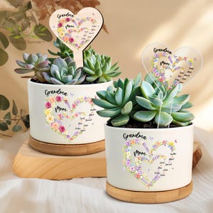 May include: Two white ceramic planters with succulents, each featuring a heart-shaped floral design with the words "Grandma Mom." One planter has a wooden heart-shaped stake. The planters sit on wooden and bamboo bases.