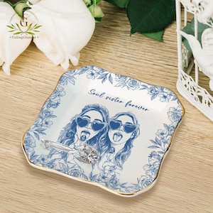 May include: A white ceramic trinket dish with a blue floral border and gold trim. The dish features an illustration of two women wearing heart-shaped sunglasses and the text "Soul sister forever". A silver ring with a large gemstone rests on the dish.