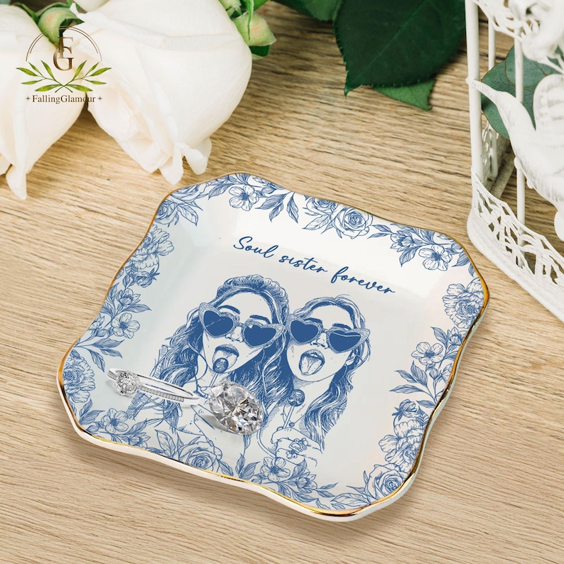 May include: A white ceramic trinket dish with a blue floral border and gold trim. The dish features an illustration of two women wearing heart-shaped sunglasses and the text "Soul sister forever". A silver ring with a large gemstone rests on the dish.