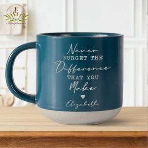 May include: Teal blue ceramic mug with a white base. The mug has a white inscription that reads "Never forget the difference that you make" with a small heart and the name "Elizabeth" below.