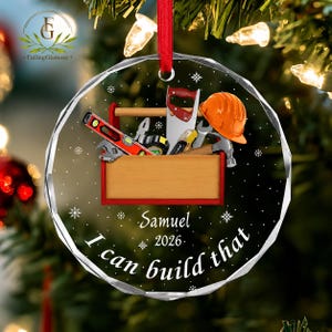 May include: A round, clear glass Christmas ornament featuring a wooden toolbox graphic filled with tools, including a level, saw, and orange hard hat. The text reads Samuel, 2026, and I can build that, surrounded by white snowflake accents.