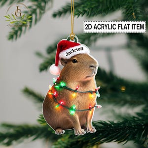 May include: A brown capybara Christmas ornament wearing a Santa hat with the name "Jackson" and wrapped in colorful Christmas lights. The ornament is a 2D acrylic flat item.